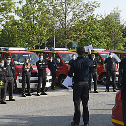 Feuerwehrleute in schwarzer Uniform und Mundschutz stehen vor Feuerwehrfahrzeugen bei einer Masken-Verteilaktion im Freien
